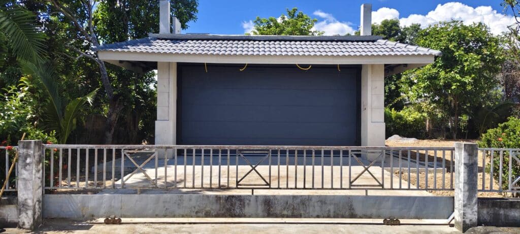 ประตูโรงรถสีเทาเข้มใต้หลังคากระเบื้องและรั้วด้านหน้า